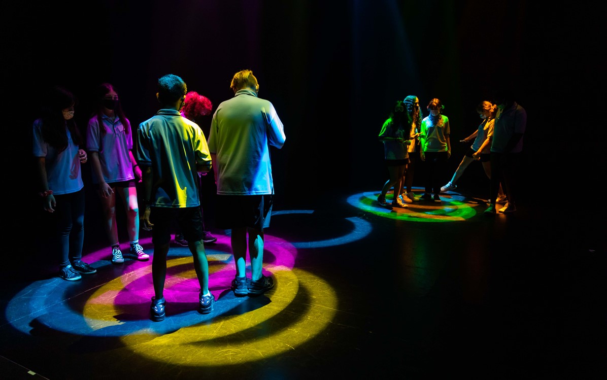Young people are standing and walking through colourful light projections in a dark space in groups of three or four